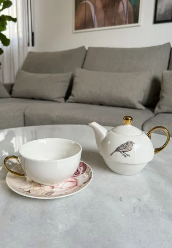 Juego de té individual con tetera, taza y platillo sobre una mesa de mármol, delante de un sofá gris